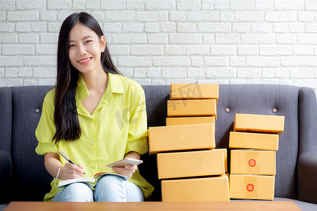 订单查看摄影照片_年轻的亚洲女性是企业家，他们在家里用笔记本电脑在线查看电话和写客户订单、网上购物、物流和送货，女性是在沙发上工作的卖家，商务中小企业。
