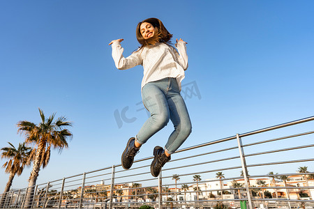 移动空调排风管摄影照片_快乐的微笑女孩高高地跳到户外，双臂高高地低头看着镜头，头发被风吹乱。