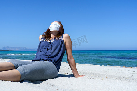 冠状病毒海滨假期：一名妇女坐在沙滩上的沙滩上，戴着 Covid-19 大流行病的面具看着太阳