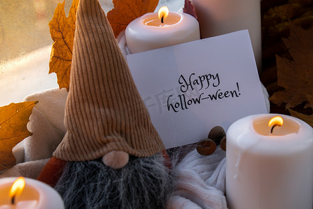 HAPPY HOLLOWEEN 文字贺卡概念 在窗台上舒适的家中庆祝万圣节秋季假期 Hygge 审美氛围 秋叶侏儒香料和蜡烛在温暖的黄色灯光下的针织白色毛衣上
