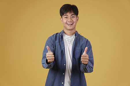 好帅的表情摄影照片_面带微笑、积极向上的亚洲年轻男子的肖像，穿着随意，大拇指竖起，赞同黄色背景中孤立的表情