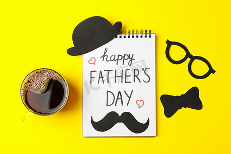 父亲节文字背景摄影照片_笔记本上刻有父亲节快乐、咖啡、装饰性领结、眼镜、胡须和彩色背景上的帽子、文字空间