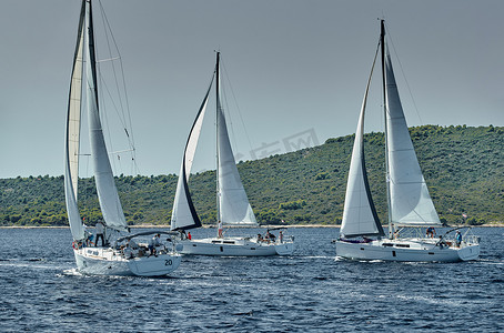 帆摄影照片_帆船在日落时参加帆船赛，帆船比赛，水面上的帆反射，五颜六色的大三角帆，船尾的船数，岛屿在背景中，天气晴朗