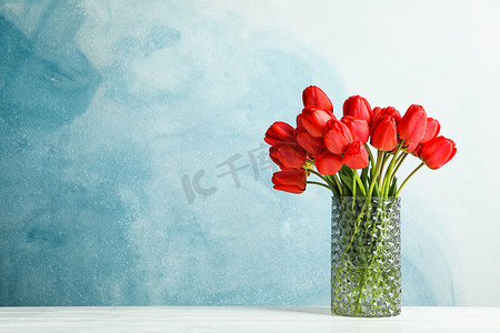 红色文字背景摄影照片_蓝色背景的白桌上有美丽红色郁金香的玻璃花瓶，文字空间