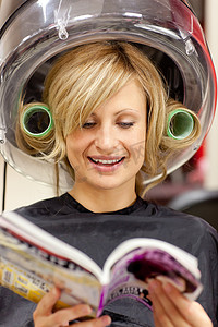创意杂志摄影照片_快乐的女人在吹风机下阅读一本带卷发器的杂志