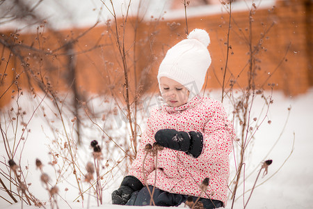 一个小女孩坐在雪地里，戴着连指手套采摘干植物