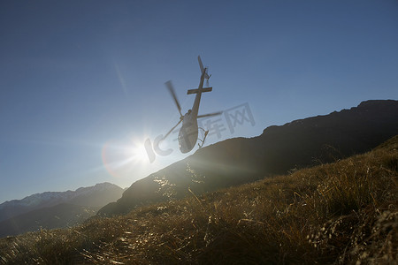 蓝色的直升飞机摄影照片_一架直升飞机在太阳前飞过山丘的低角度视图