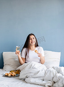 年轻的黑发女人坐在床上醒着，带着红心形气球和装饰品，喝着香槟，吃着羊角面包