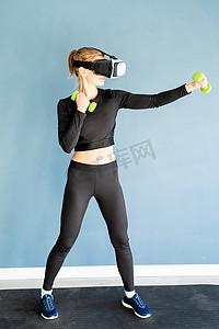 身穿运动服、戴着虚拟现实眼镜的年轻金发女性站在健身垫上，用哑铃锻炼