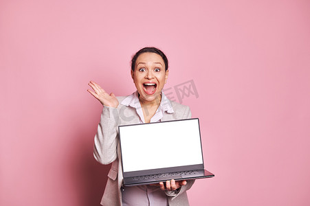 惊讶的多民族女性张着嘴，拿着带白色空白屏幕的笔记本电脑，与粉红色背景隔离