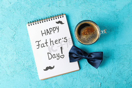 父亲节文字背景摄影照片_笔记本上印有快乐父亲节、蓝色领结和彩色背景咖啡、文字空间和顶视图