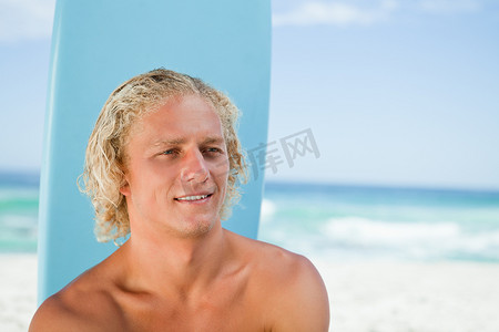 泳衣帅哥摄影照片_带着冲浪板坐在沙滩上微笑的年轻人