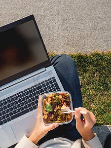 女人带着笔记本电脑坐在公园长椅上，拿走午餐盒。