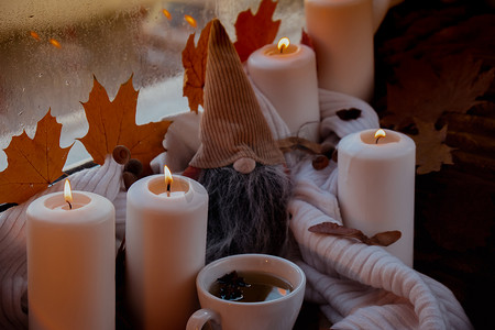 万圣节氛围摄影照片_感恩节和 Hello Fall 万圣节概念在舒适的家中在窗台上庆祝秋季假期 Hygge 审美氛围秋叶侏儒、香料和蜡烛在温暖的黄色灯光下的针织白色毛衣上。