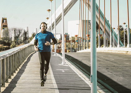 奔跑广告摄影照片_穿着蓝色衬衫的年轻人在桥上奔跑，戴着耳机听音乐。