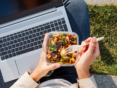 女人带着笔记本电脑坐在公园长椅上，拿走午餐盒。