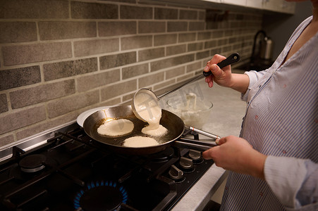 一位女厨师、家庭主妇的特写镜头，将圆形面糊倒入黑色炉子上的煎锅中，并为忏悔节准备煎饼。