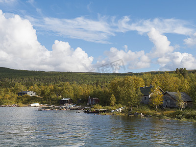 从 Lulealven 河看 Saltoluokta 的萨米小屋村，那里有青山、黄桦树和蓝天白云，黄金时段的灯光
