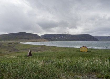 在冰岛西峡湾自然保护区 Hornstrandir 的阿达尔维克湾 latrar 营地和黄色紧急避难所的景色，有绿草草甸、石滩、海洋、积雪覆盖的山丘和乌云背景