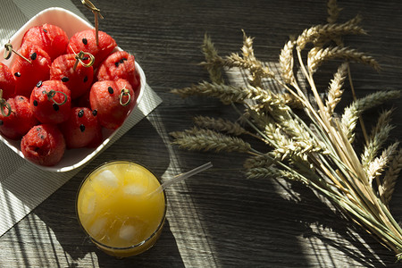 红色穗摄影照片_阳光下木桌上的鲜榨橙汁、甜西瓜甜点和成熟小麦的小穗……