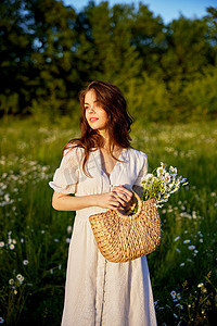 穿着浅色连衣裙的可爱女人站在田野里微笑着，手里拿着一篮子雏菊