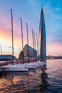 俄罗斯，圣彼得堡，2020 年 7 月 3 日：日落时的拉赫塔中心摩天大楼，玻璃幕墙中日落天空的倒影，它是欧洲最高的摩天大楼，摩天大楼港口的帆船