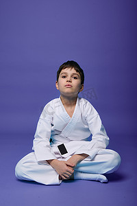 儿童跆拳道摄影照片_自信的欧洲少年 — 合气道斗士 — 身着白色和服，坐在莲花姿势中，同时在紫色背景和复制空间中练习东方武术