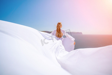 一位长发的金发女子身着飘逸的白色长裙站在阳光明媚的海边，丝绸面料随风飘扬，背景是蓝天和群山。