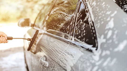 在自助洗车中清洗的银色汽车的侧面，人手拿着的刷子在洗发水中留下笔触，背景是阳光背光。