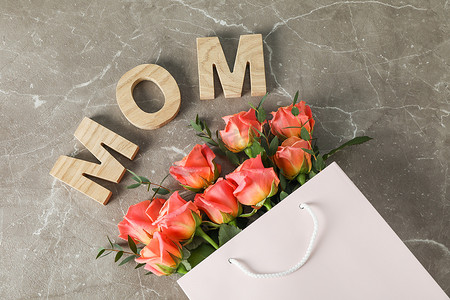 花束妈妈摄影照片_带橙色玫瑰花束的礼品袋和棕色背景上的妈妈铭文，顶视图