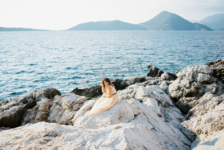 新娘坐在多岩石的海边，背靠群山，手托着头
