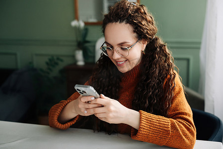 戴着眼镜、微笑的年轻女性拿着智能手机，使用手机现代技术，看着手机，检查手机应用程序，发短信和浏览互联网，坐在家里购物