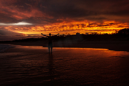 年轻人在空中举手，因为他很高兴，背景是澳大利亚阳光海岸努萨维尔海滩的彩色日落。