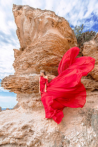 一位身着红色丝绸连衣裙的女子站在海边，背靠群山，她的裙子在微风中摇曳。