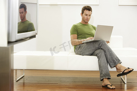 现代多人沙发摄影照片_在现代公寓的沙发上使用笔记本电脑的全长年轻人