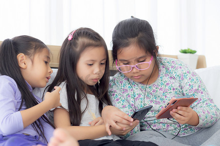 儿童智能摄影照片_快乐的亚洲小孩和朋友在家里的沙发上玩智能手机，孩子们在沙发上一起使用手机，女孩看智能手机以获得娱乐、生活方式和沟通理念。
