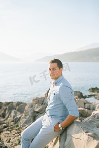 新郎坐在海边的岩石海岸上，双手插在口袋里