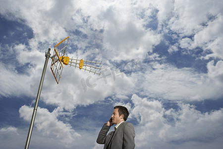 一加手机摄影照片_一位年轻商人在卫星塔下和多云天空下使用手机的低视角