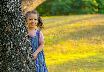 亚洲小女孩呆在大树后面，在花园里带着晨光微笑。