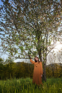 一位长着红色长发的甜美迷人的女人穿着橙色长裙，站在一棵开花的树旁，用手抚摸着树枝，看着镜头