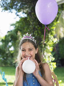 气球吹气摄影照片_在生日聚会上戴着头饰和吹气球的微笑小女孩的画像