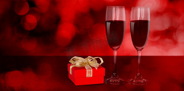 桌子上有两个酒杯和红色礼盒，红色模糊散景背景、香槟酒杯和带有庆祝和周年纪​​念、爱情和浪漫、情人节概念的礼物。