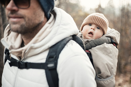 运动型父亲背着他的小儿子穿着冬季连身衣和帽子，背着背包在秋天的森林里徒步旅行。