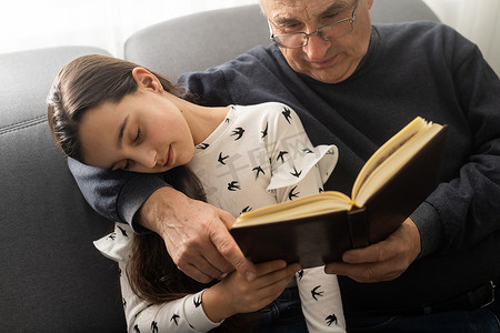 老爷爷小漂亮孙女坐在沙发上拥抱住家检疫安全的照片，在室内阅读有趣的童话故事现代室内客厅.