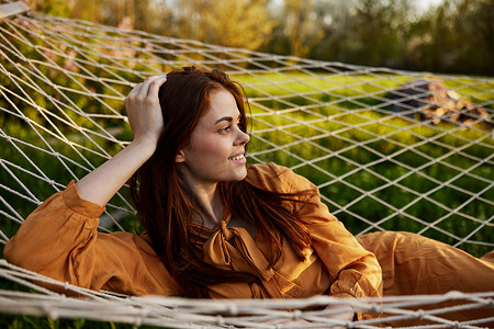 一个快乐的女人躺在网状吊床上休息，头枕在手上，微笑着幸福地看着别处，在夕阳的余晖中享受温暖的一天，躺在橙色的裙子里