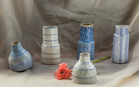 粉色花朵装饰摄影照片_五个蓝色和白色手工陶瓷花瓶，粉色花朵装饰在腮红纹理桌布上。