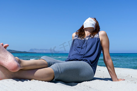 感染摄影照片_冠状病毒海滨假期：一名妇女坐在沙滩上的沙滩上，戴着 Covid-19 大流行病的面具看着太阳