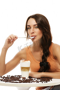 嘴里叼着勺子的漂亮女人坐在桌边，手里拿着拿铁玛奇朵咖啡