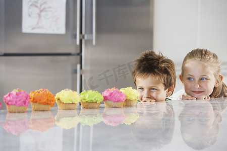 小女孩和男孩从柜台上偷看一排纸杯蛋糕