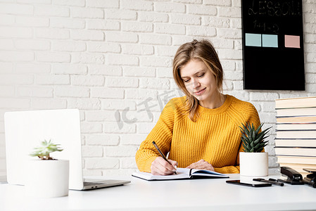 穿着黄色毛衣的年轻微笑女性使用笔记本电脑在笔记本上写作在线学习